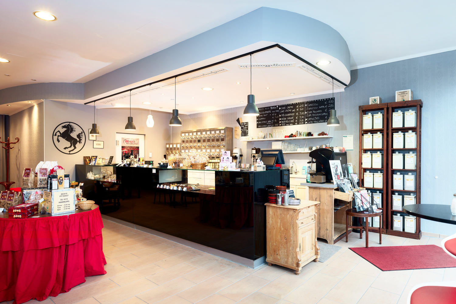 Große, glänzend schwarze Verkaufstheke mit Pralinen, Kaffeeschütten und großer Espressomaschine. Teeregale und ein roter Warentisch sowie unser Stuttgarter Rössle als Edelstahlbild